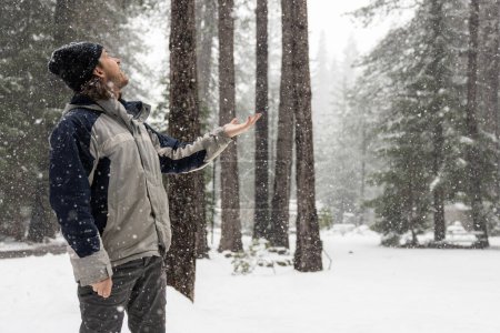 Téléchargez les photos : Un homme vêtu d'une veste grise et bleue se tient debout dans la neige, tenant la main pour attraper des flocons de neige. Concept d'émerveillement et d'appréciation de la beauté du paysage hivernal - en image libre de droit