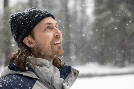 Téléchargez les photos : Un homme portant un chapeau noir et une veste grise sourit en regardant la neige tomber du ciel - en image libre de droit