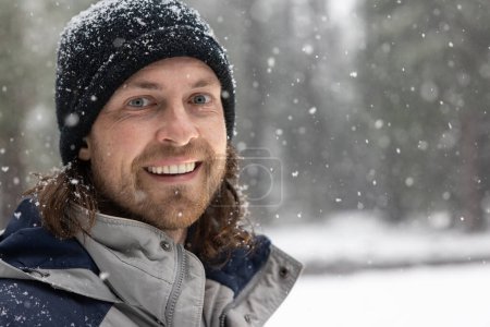Téléchargez les photos : Un homme avec un chapeau noir et une barbe sourit dans la neige. Il porte une veste bleue et une écharpe grise - en image libre de droit