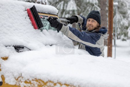 Foto de Un hombre está de pie en la nieve, sosteniendo un cepillo de nieve y sonriendo. Está limpiando el parabrisas de un jeep amarillo. - Imagen libre de derechos