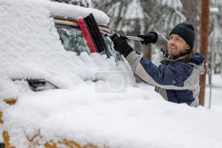 Foto de Un hombre está limpiando el parabrisas de un coche con un cepillo de nieve. El hombre sonríe y disfruta de la tarea - Imagen libre de derechos