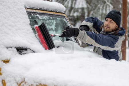Foto de Un hombre está limpiando la nieve de la ventana de un coche. La escena es fría y nevada, y el hombre lleva un sombrero negro. - Imagen libre de derechos