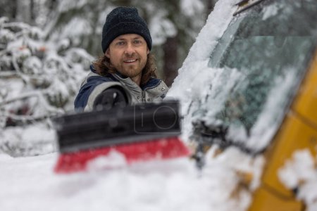 Téléchargez les photos : Un homme sourit alors qu'il pousse la neige d'une voiture. La scène se déroule dans un environnement enneigé, et l'homme porte un chapeau noir - en image libre de droit