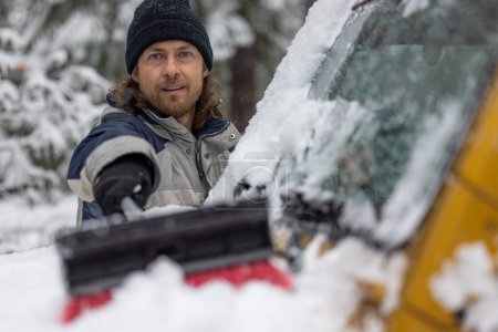 Téléchargez les photos : Un homme nettoie la neige d'une voiture avec une brosse à neige. L'homme porte un chapeau noir et une veste bleue. - en image libre de droit