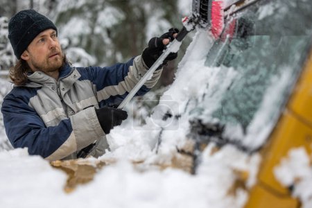 Téléchargez les photos : Un homme avec un chapeau noir nettoie la neige d'un pare-brise de voiture. La scène se déroule dans un environnement enneigé, et l'homme est concentré sur sa tâche - en image libre de droit