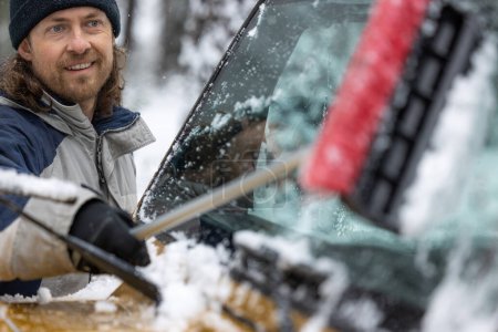 Foto de Un hombre está limpiando el parabrisas de su coche con un cepillo de nieve. Él sonríe y disfruta de la tarea - Imagen libre de derechos