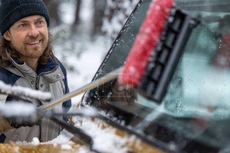 Téléchargez les photos : Un homme nettoie le pare-brise de sa voiture avec une brosse à neige. Il sourit et il apprécie la tâche - en image libre de droit