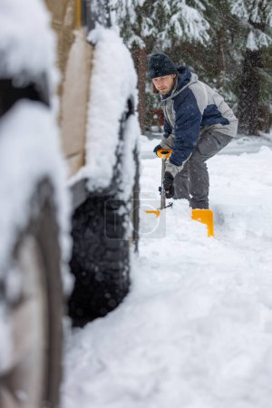 Téléchargez les photos : Un homme vêtu d'une veste bleue et grise pelle la neige du sol. Il porte des bottes jaunes et un chapeau noir. - en image libre de droit