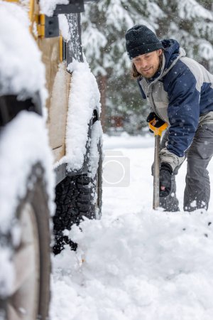Téléchargez les photos : Un homme vêtu d'un chapeau noir et d'une veste bleue pelle la neige sur le dos d'une jeep jaune - en image libre de droit