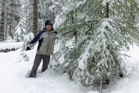 Téléchargez les photos : Un homme est debout dans la neige à côté d'un arbre. Il porte un chapeau noir et une veste bleue. - en image libre de droit