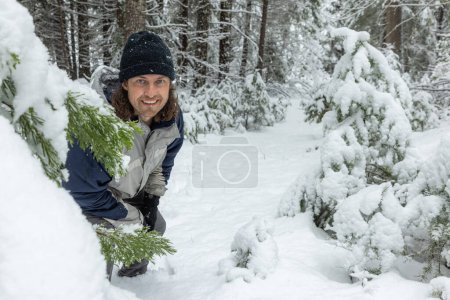 Ein Mann steht im Schnee und blickt auf einen Baum. Er trägt eine schwarze Mütze und eine blaue Jacke. Konzept von Abenteuer und Erkundung, wie der Mann in einem verschneiten Wald ist, umgeben von Bäumen