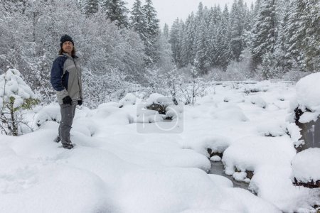 Téléchargez les photos : Un homme se tient dans la neige, les mains dans les poches. La neige est profonde et les arbres sont nus. Il porte une veste bleue et un chapeau. La scène est calme et paisible - en image libre de droit