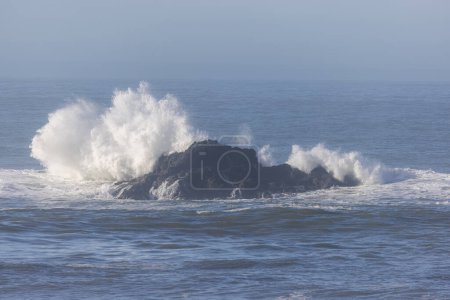 Foto de Una gran roca está en el océano con una ola que se estrella sobre ella. El agua está tranquila y el cielo azul - Imagen libre de derechos