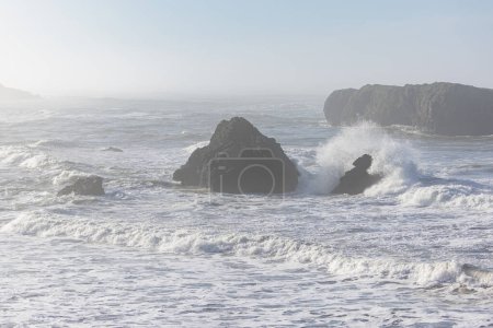 Foto de El océano está tranquilo y las olas son pequeñas. Las rocas están esparcidas por el agua. El cielo está despejado y el sol brilla - Imagen libre de derechos