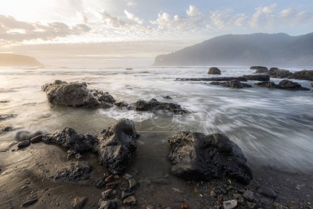 Foto de El océano está tranquilo y las rocas están dispersas a lo largo de la orilla. El cielo está nublado y el sol se pone - Imagen libre de derechos