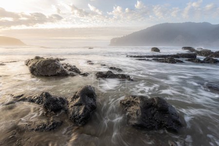 Téléchargez les photos : L'océan est calme et les rochers sont éparpillés dans l'eau. Le ciel est nuageux et le soleil se couche - en image libre de droit
