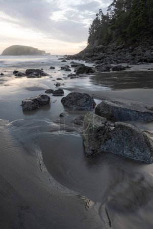 Téléchargez les photos : Une plage rocheuse avec un rivage rocheux et un plan d'eau en arrière-plan. La plage est principalement couverte de rochers, avec un peu de sable éparpillé autour. Le ciel est nuageux et le soleil se couche - en image libre de droit