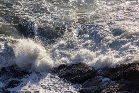 Téléchargez les photos : Une grosse vague s'écrase contre un rivage rocheux. L'eau est blanche et mousseuse, et les rochers sont éparpillés dans toute la scène. La scène est puissante et dramatique - en image libre de droit