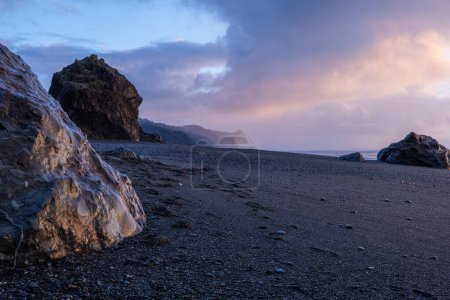 Téléchargez les photos : Une plage rocheuse avec un grand rocher au premier plan. Le ciel est nuageux et le soleil se couche - en image libre de droit