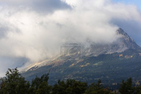 Téléchargez les photos : Une montagne couverte de nuages avec quelques arbres au premier plan. Le ciel est nuageux et la montagne est couverte de neige - en image libre de droit