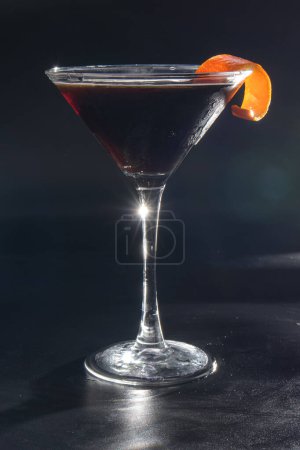 Foto de Una copa de martini con una rebanada de cáscara de naranja en la parte superior. El vaso está lleno de un líquido oscuro - Imagen libre de derechos