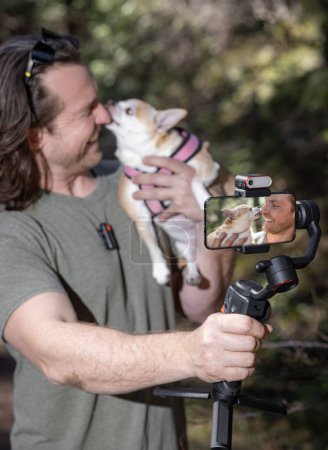 Téléchargez les photos : Un homme utilise une carabine pour se filmer lui-même et un chihuahua qu'il tient. Le film se déroule dans une forêt naturelle. - en image libre de droit