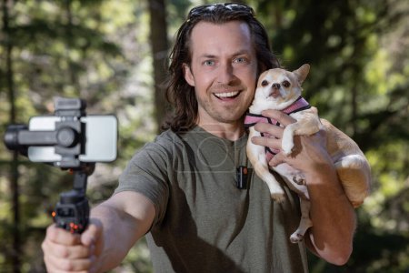 Foto de Un hombre que usa un cardán telefónico para grabarse a sí mismo y a un chihuahua que sostiene. La película está ocurriendo en un lugar de bosque natural. - Imagen libre de derechos