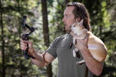 Man using a phone gimbal to film himself and a chihuahua he is holding. The film is happening in a natural forest location.
