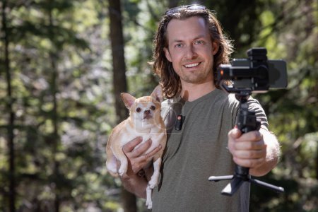 Téléchargez les photos : Un homme utilise une carabine pour se filmer lui-même et un chihuahua qu'il tient. Le film se déroule dans une forêt naturelle. - en image libre de droit
