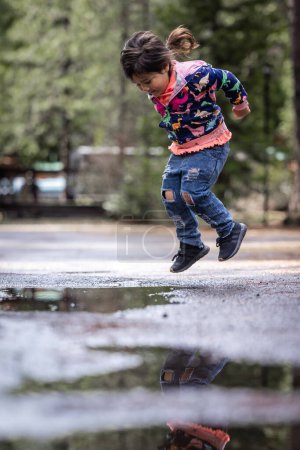 Téléchargez les photos : Une jeune fille saute dans une flaque d'eau. Concept de joie et de jeu, comme la fille s'amuse tout en éclaboussant dans l'eau. La flaque reflète l'environnement environnant - en image libre de droit
