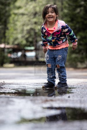 Foto de Una joven está de pie en un charco de agua, vistiendo una chaqueta azul y rosa. Ella sonríe y se divierte. - Imagen libre de derechos
