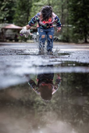 Téléchargez les photos : Une jeune fille joue sous la pluie, éclaboussant l'eau et tenant un animal en peluche. La scène est ludique et joyeuse, avec la fille profitant du moment et de l'environnement humide - en image libre de droit