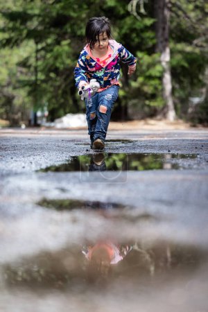 Foto de Una joven está caminando por un camino mojado, con su reflejo en el agua. La escena es tranquila y serena, con la chica disfrutando de su tiempo al aire libre - Imagen libre de derechos