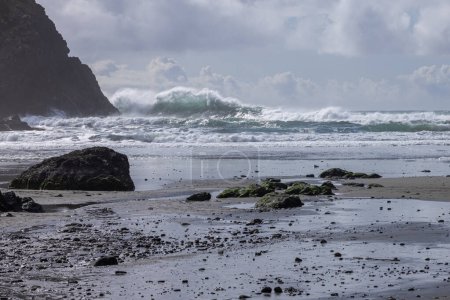 Foto de Una playa rocosa con una gran ola chocando contra la orilla. El agua está agitada y el cielo nublado - Imagen libre de derechos