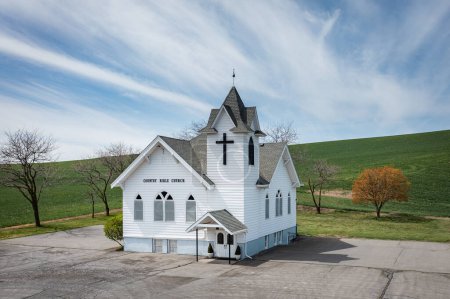 Photo for A small white church with a cross on top. The church is surrounded by trees and grass. The sky is blue and there are clouds in the background - Royalty Free Image