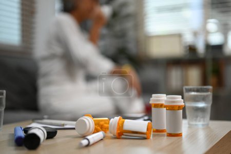 Focus on pills bottles and glass of water with senior woman sitting in background. Medicine, pharmacology, healthcare concept.