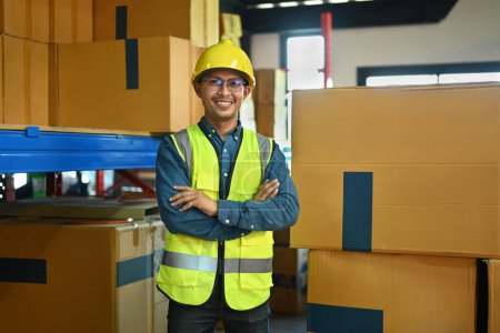Portrait of smiling male manager wearing hardhat and reflective jacket standing with arms crossed between shelves with cardboard boxes.