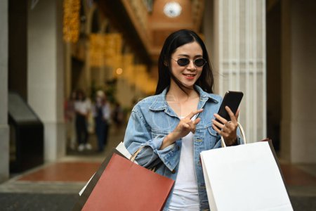 Photo for Beautiful fashionable asian woman carrying shopping bags and using smart phone outdoor front of the shopping center. - Royalty Free Image