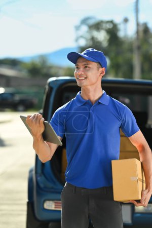 Foto de Retrato del repartidor vistiendo uniforme azul con cartón en las manos, buscando la dirección en la tableta digital. - Imagen libre de derechos