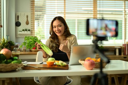 Foto de Nutricionista femenina confiada sosteniendo apio, proporcionando consultas en línea o grabando contenido de video en un teléfono inteligente. - Imagen libre de derechos