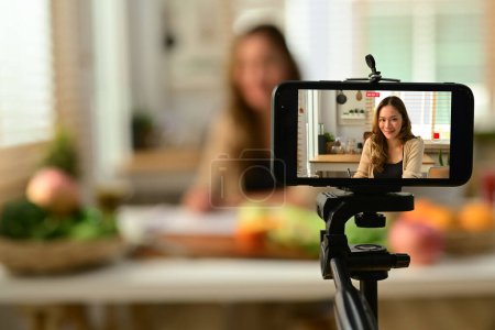 Una bloguera confiada sosteniendo apio, proporcionando consultas en línea o grabando contenido de video en un teléfono inteligente.