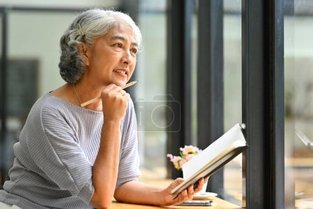 Foto de Hermosa mujer madura de pelo gris sosteniendo libro y mirando hacia otro lado, soñando despierto o pensando en buenos planes de jubilación. - Imagen libre de derechos