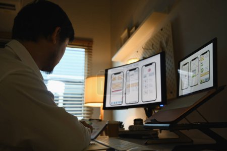 Foto de Joven diseñador que trabaja en la computadora personal con diseño de aplicaciones móviles en la pantalla en la habitación oscura. - Imagen libre de derechos