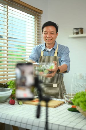Foto de Alegre macho senior mostrando tazón de ensalada frente al teléfono inteligente durante la grabación de vídeo vlog en la cocina - Imagen libre de derechos