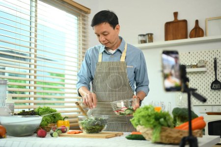Foto de Hombre de mediana edad grabación de vídeo con teléfono inteligente sobre la sesión tutorial de ensalada vegana saludable en la cocina - Imagen libre de derechos
