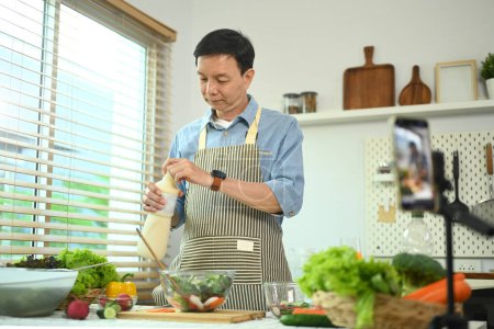 Foto de Hombre de mediana edad activa grabación de video blog o vlog sobre alimentos saludables con teléfono inteligente en la cocina casera. - Imagen libre de derechos