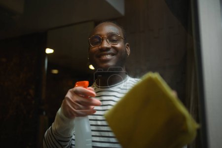 Foto de Sonriente joven africano con guante de goma limpiando espejo en el baño con trapo. - Imagen libre de derechos
