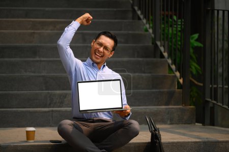 Foto de Hombre de negocios alegre sosteniendo portátil con pantalla en blanco y levantando la mano en sí gesto. - Imagen libre de derechos