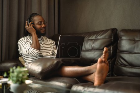 Foto de Hombre africano relajado en auriculares con portátil en el sofá y escuchar música con los ojos cerrados. - Imagen libre de derechos