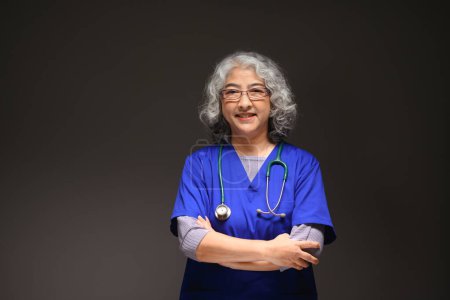 Foto de Retrato de amigable doctora madura con estetoscopio alrededor de su cuello de pie sobre fondo negro. - Imagen libre de derechos